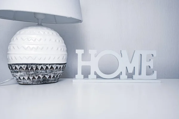 Керамическая лампа у кровати с текстильной абажуром на белом столе с табличкой дома, уютная концепция дома в серых пастельных тонах — стоковое фото