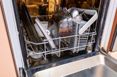 Open overloaded dishwasher with different dishes and kitchen utensils