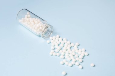 Homeopathic globules scattered from a clear glass bottle on light blue background, alternative homeopathy medicine concept.