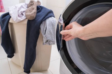 Çamaşır sepetinde yatan kirli çamaşırları yıkamak için çamaşır makinesinin kapısını açan bir kadın..