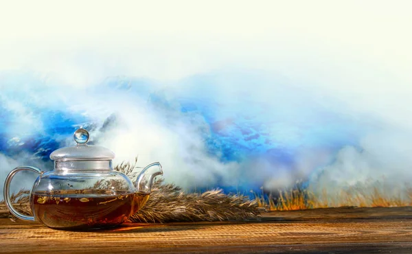 glass transparent teapot with hot tea on a wooden old background and twigs of reeds on a background of mountains and clouds