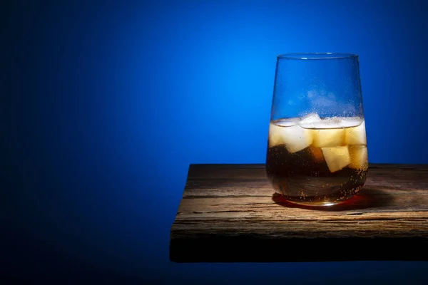 Glass glass with ice cubes and carbonated drink standing on a wooden table on a gradient dark background