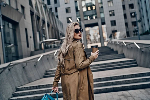 オフィスビル近くの街で屋外を歩きながらコーヒー カップを保持しているファッショナブルな魅力的な金髪女性 — ストック写真