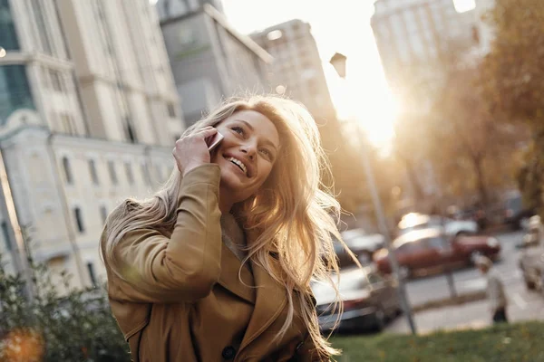スマート フォンで屋外で立って話す魅力的な金髪の若い女性 — ストック写真