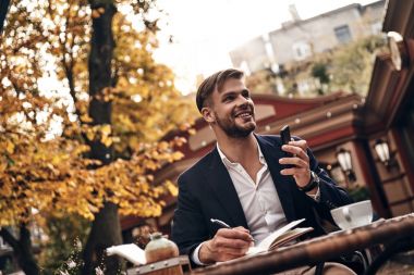 genç adam akıllı casual giyim akıllı telefon tutarak ve açık havada restoranda otururken gülümseyen