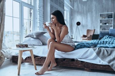 esmer genç kadın yatakta evde otururken kahve tutan pijama