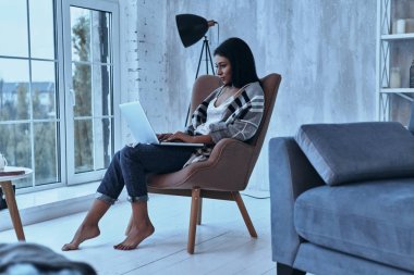 Evde koltukta otururken dizüstü bilgisayar kullanan çekici blogger kadın