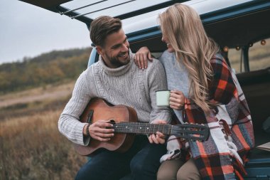 Yakışıklı genç gitar çalmak onun güzel kız arkadaşı için mavi retro tarzı mini van bagajında otururken adam
