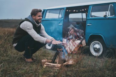 man warming up by the bonfire while woman reading a book in car transporter