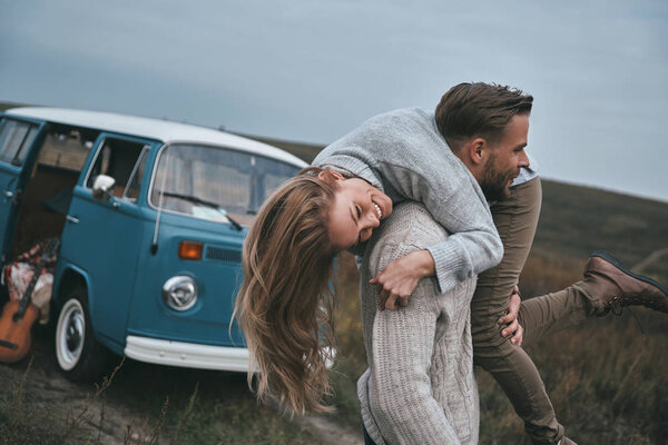 man carrying his happy smiling girlfriend on shoulders near blue retro style mini van car