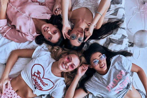 Top view of four playful young women in pajamas and eyewear lying on bed at home