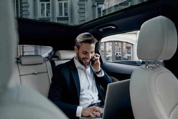 Bonito Jovem Terno Completo Falando Telefone Inteligente Usando Laptop Carro — Fotografia de Stock