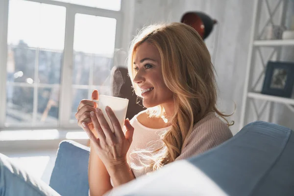 Attractive blonde woman holding cup and looking away with smile while sitting on sofa at home