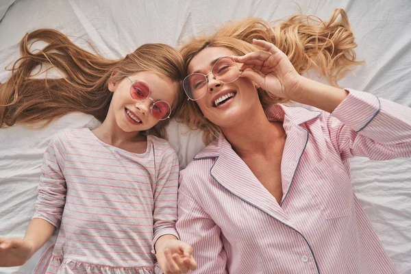 年轻漂亮的母亲和可爱的女儿戴着眼镜 躺在床上面带微笑的顶部视图 — 图库照片