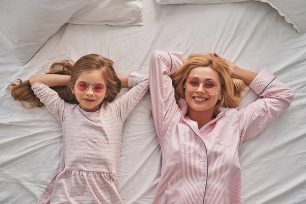 Top view of beautiful mother and cute daughter in sun glasses keeping hands behind head and smiling while lying on bed