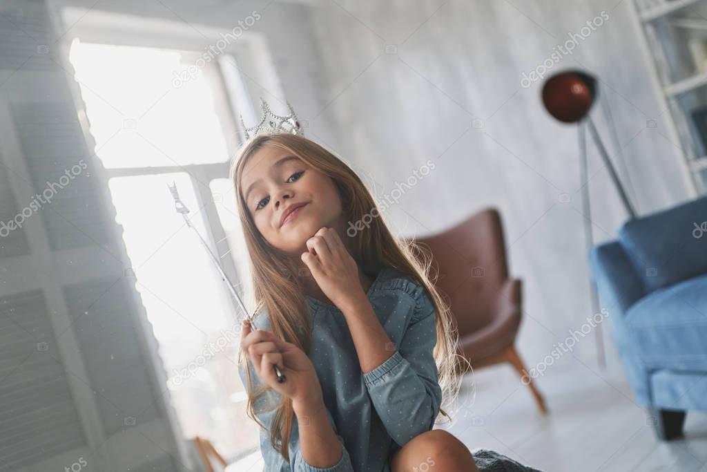 Prideful little girl keeping hand on chin and playing with a magic wand at home