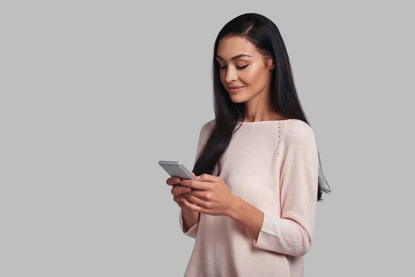 黑发妇女使用智能手机站立反对灰色背景 — 图库照片