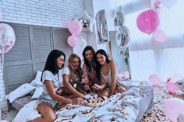 Dört güzel genç kadın pijama yatak balonlar ve konfeti ile pijama partisi yaparken pasta yemeye gülümseyen