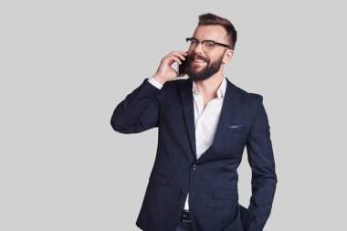 Yakışıklı genç adam akıllı telefonda konuşuyor ve gri arka plan karşı dururken gülümseyen resmi kıyafeti