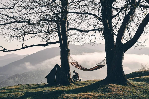 autumn camping in mountains, hanging hammock on trees, man sitting on background