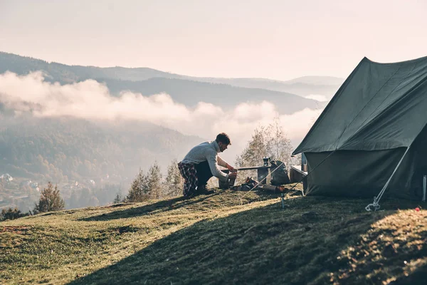 man making cooking on campfire while sitting near tent in mountains