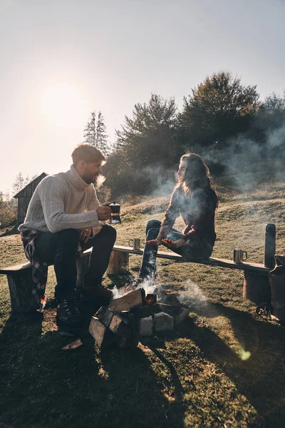 焚き火の近くの山で屈託のない時間を過ごす中にホットド リンクを持つ愛のカップル — ストック写真