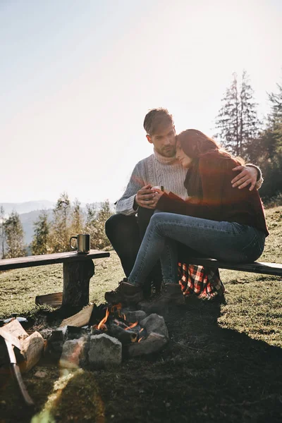 白人情侣坐在长凳附近的篝火在山上的热饮料杯 — 图库照片