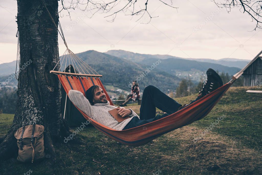 smiling man lying in hammock with book while his girlfriend enjoying beauty in nature, autumn camping in mountains