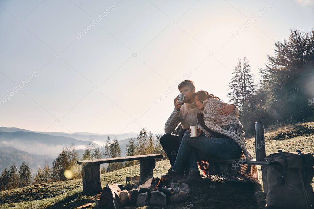 couple in love having hot drinks while spending carefree time in mountains near bonfire