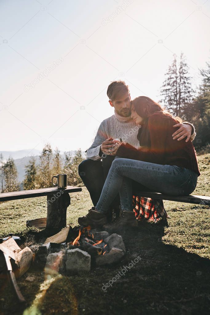 Caucasian couple in love sitting on bench near bonfire in mountains with hot drinks in cups