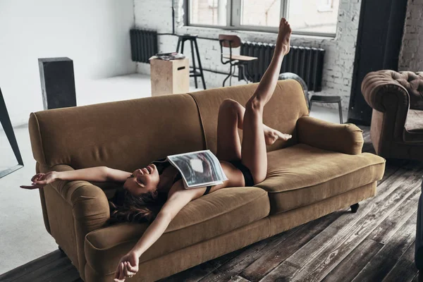 怠惰な女性が雑誌を笑顔で覆われて 楽しみながら自宅のソファーに横になっていると — ストック写真
