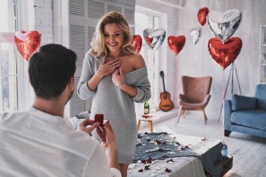 blonde young woman looking at engagement ring with smile while her boyfriend proposing her to marry clipart