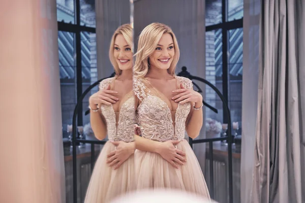 Reflection of beautiful young woman wearing wedding dress and smiling in front of mirror in bridal shop