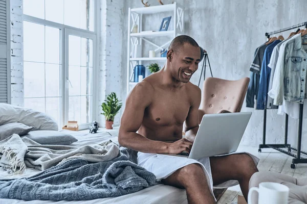 Handsome young African man covered with shower towel smiling and using laptop while sitting on bed at home