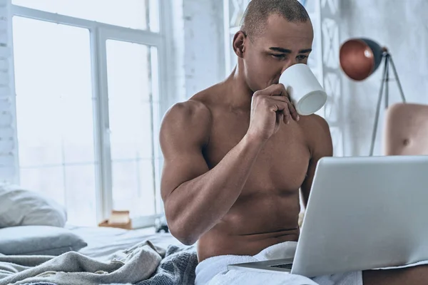 上半身裸のハンサムな若いアフリカ人コーヒーを飲むと 自宅のベッドに座ってラップトップを使用して — ストック写真