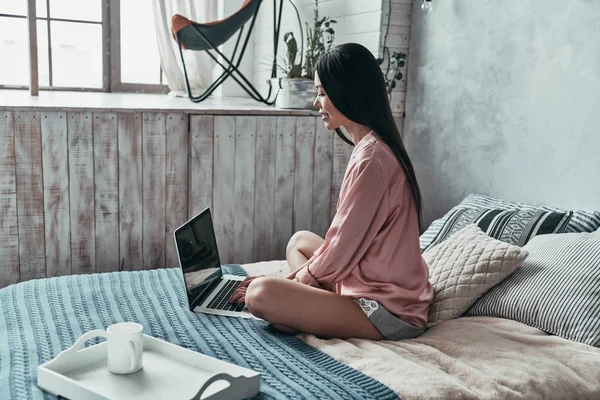 コンピューターを使用して 自宅のベッドに座って笑顔の魅力的な若い女性 — ストック写真