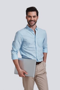 dizüstü bilgisayar taşıma ve gri arka plan karşı dururken gülümseme ile kameraya bakarak genç adam gülümseyen