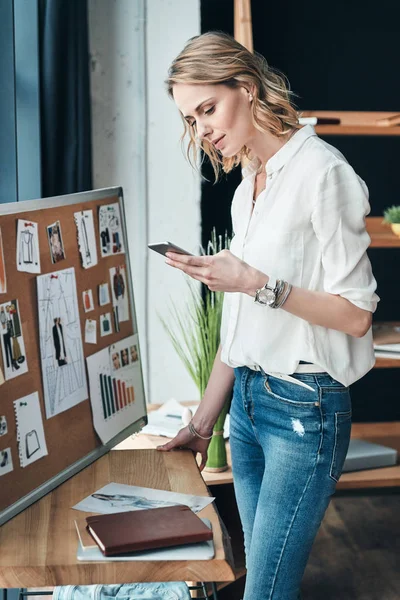 blonde woman typing business message on smart phone and standing in workshop