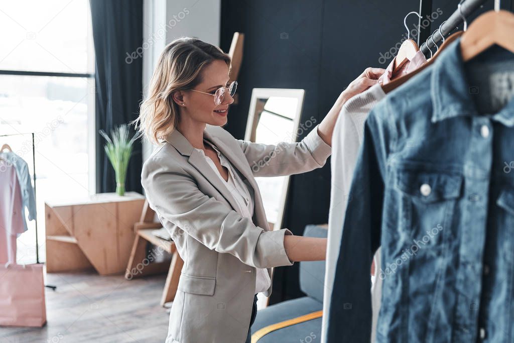 smiling blonde woman choosing clothes from rank while standing in workshop
