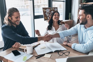 Başka bir proje bitti. Genç modern iş arkadaşları el sıkışan ve yaratıcı ofiste otururken gülümseyen akıllı rahat kıyafet