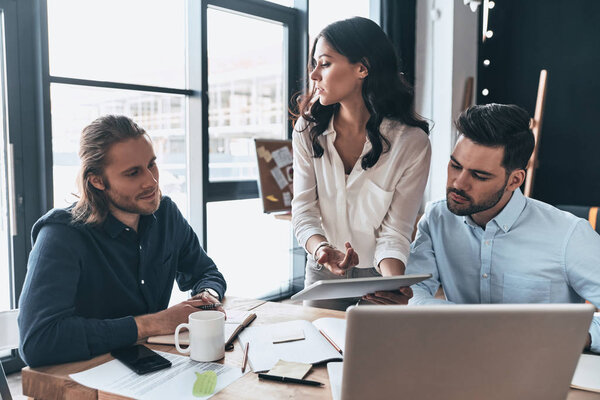 Приятный рабочий день. Молодая современная женщина в элегантной повседневной одежде держит цифровой планшет и указывает на него, работая вместе с коллегами в креативном офисе

