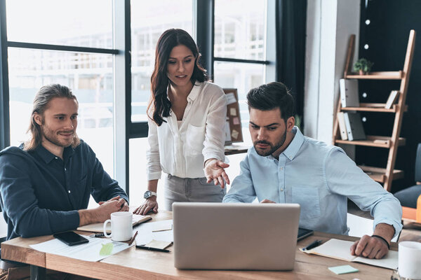 Быстрая встреча. Молодая современная женщина в элегантной повседневной одежде указывает на ноутбук и улыбается во время работы с коллегами в креативном офисе
