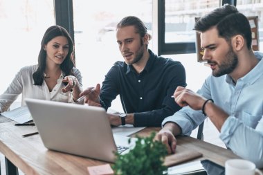 Çalışma günü. Grup içinde smart casual modern gençlerin giyim laptop işaret ve yaratıcı ofiste otururken gülümseyen