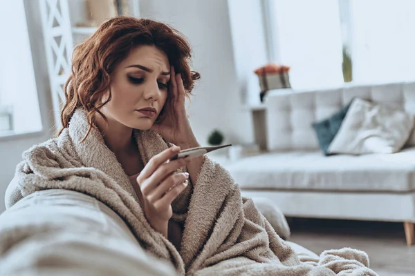 自宅のソファに腰掛けながら温度計を見て病気の若い女性 — ストック写真