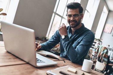 modern ofisinde çalışan ve dizüstü bilgisayar, günlük ve yazma fikirler ile masada oturan mutlu yakışıklı genç adam 