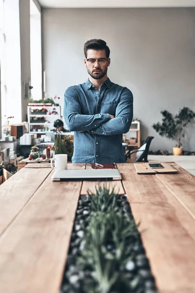 serious handsome young man looking at camera and keeping arms crossed while standing in creative working space, wooden table with plants and closed laptop
