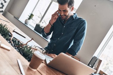 mutlu yakışıklı genç adam akıllı telefon duran tahta masada modern ofis içinde laptop ile konuşurken