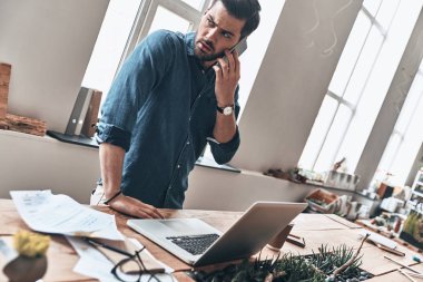 modern ofis içinde laptop ile tahta masada duran akıllı telefonda konuşurken yakışıklı genç