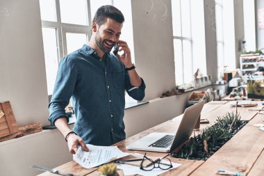 mutlu yakışıklı genç adam akıllı telefon duran tahta masada modern ofis içinde laptop ile konuşurken