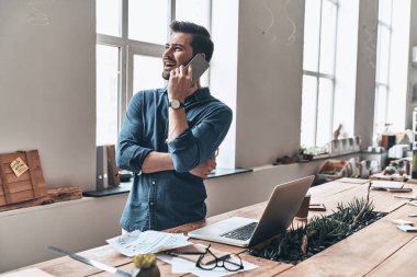 mutlu yakışıklı genç adam akıllı telefon duran tahta masada modern ofis içinde laptop ile konuşurken